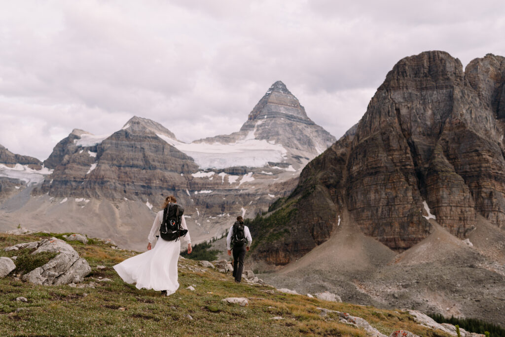 Couple with backpacks on and wedding attire walk towards Mount Assiniboine during elopement. 