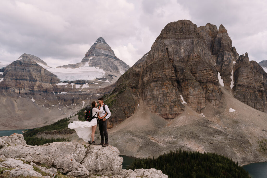 Couple stands on rock with backpacks on and dress blowing in the wind during Mount Assiniboine elopement.