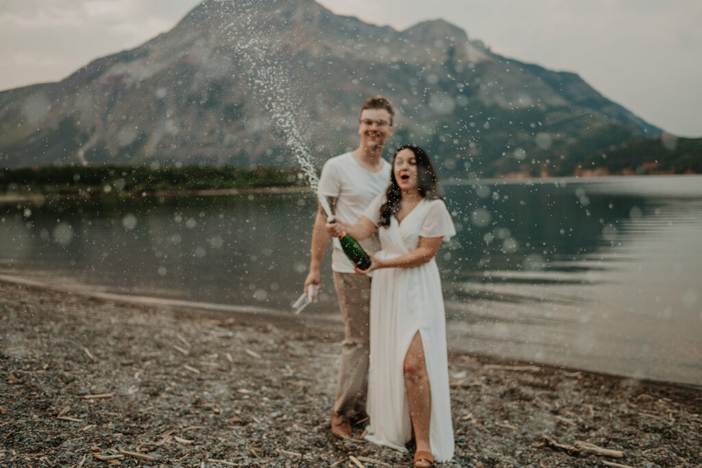 Couple pops champagne at Driftwood beach during waterton elopement