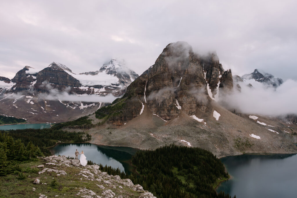 Couple stands on rock overlooking the lake and misty mountains during Mount Assiniboine Elopement