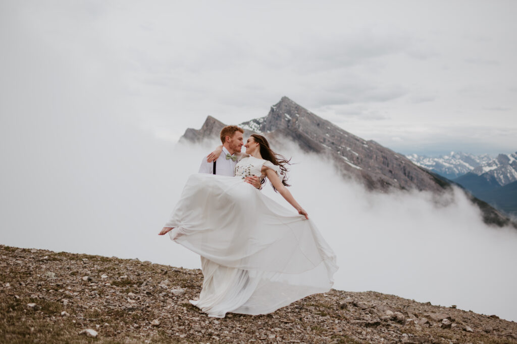 groom holds barefoot bride up while twirling her as her dress flows during a moody cloudy mountain elopement in banff national park