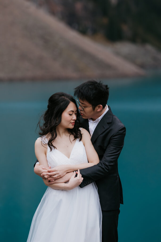 Couple snuggle up during elopement in front of bright blue water at Moraine Lake