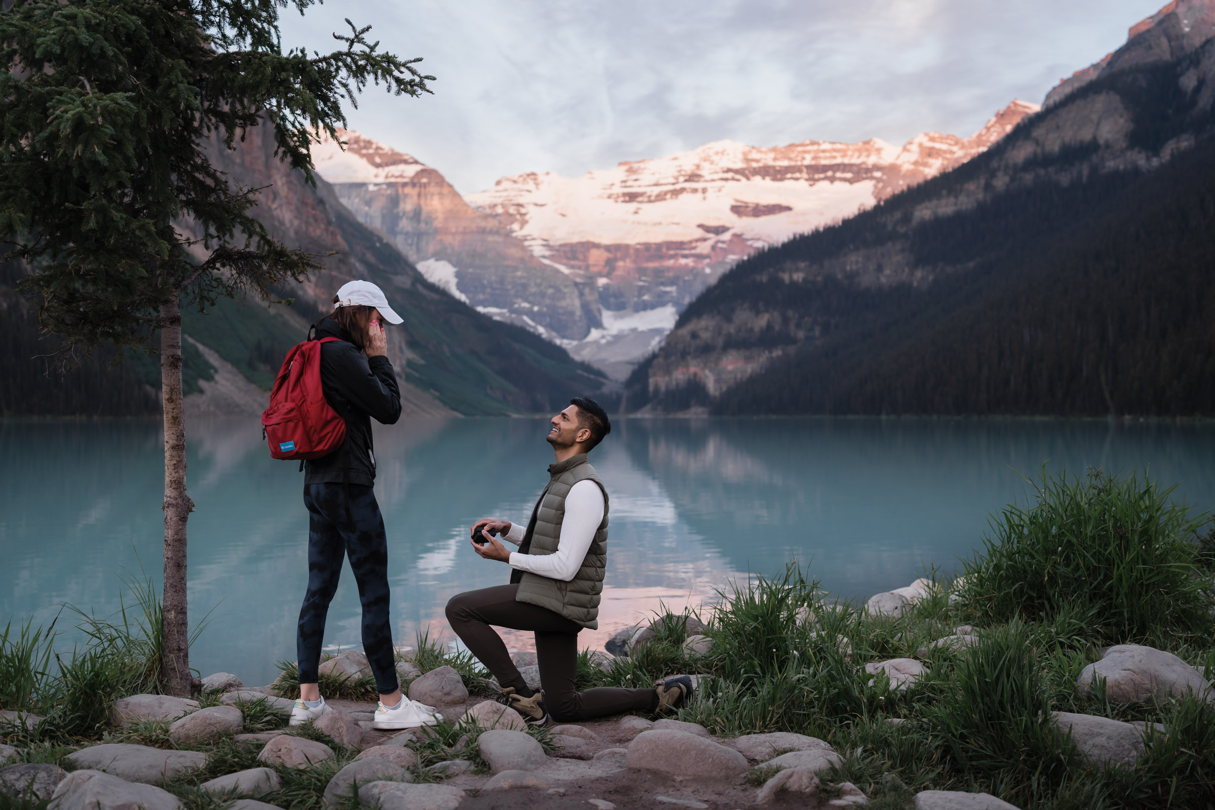Man is down on one knee infront of surprised girlfriend as the sun lights up the glacier behind Lake Louise.