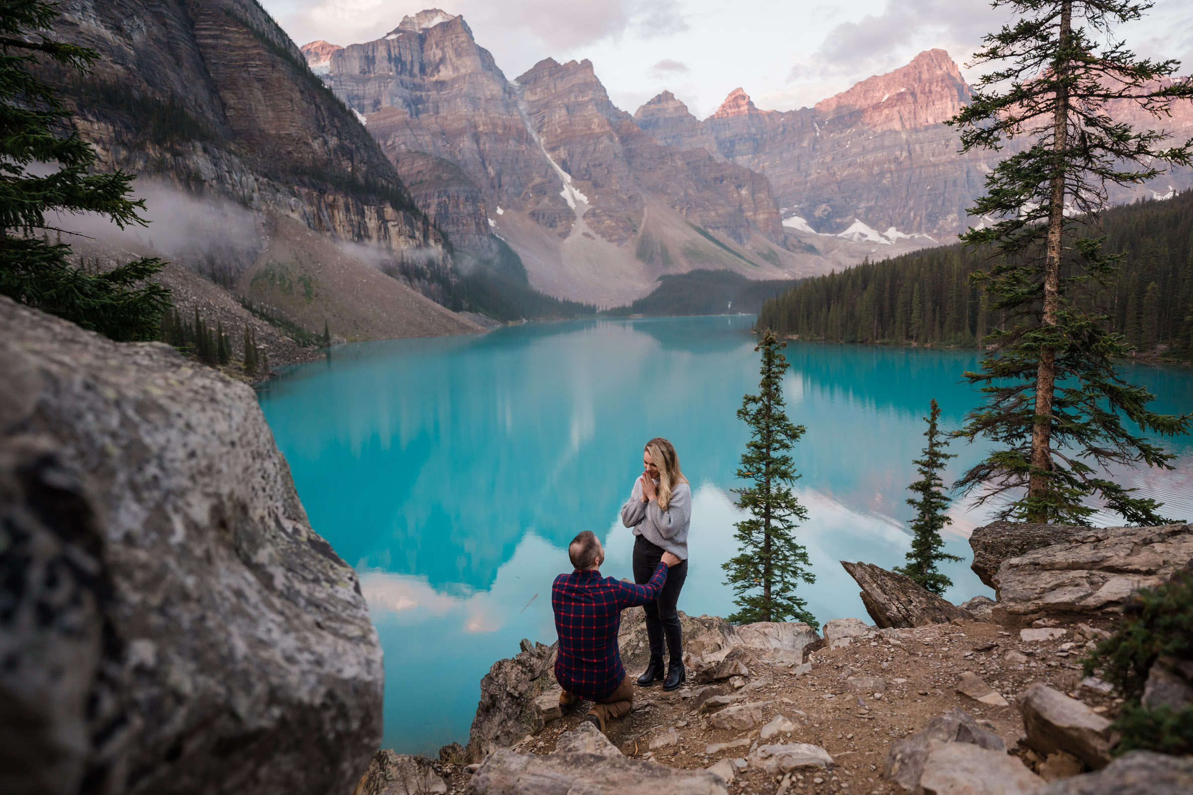 Man in plaid shirt kneels infront of his girlfriend with one hand on her hip and holding out a ring while she covers her face with her hands in surprise as he proposed during sunrise at Moraine Lake