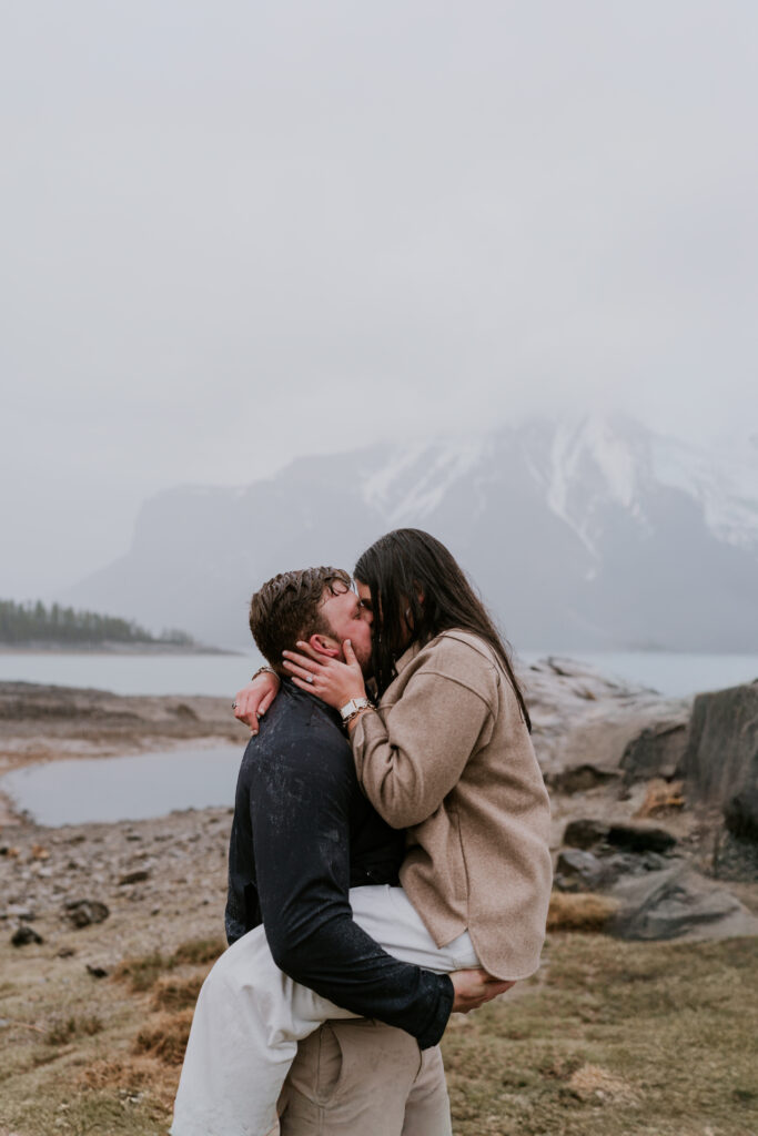 man holds woman up as they kiss as the rain falls during banff proposal