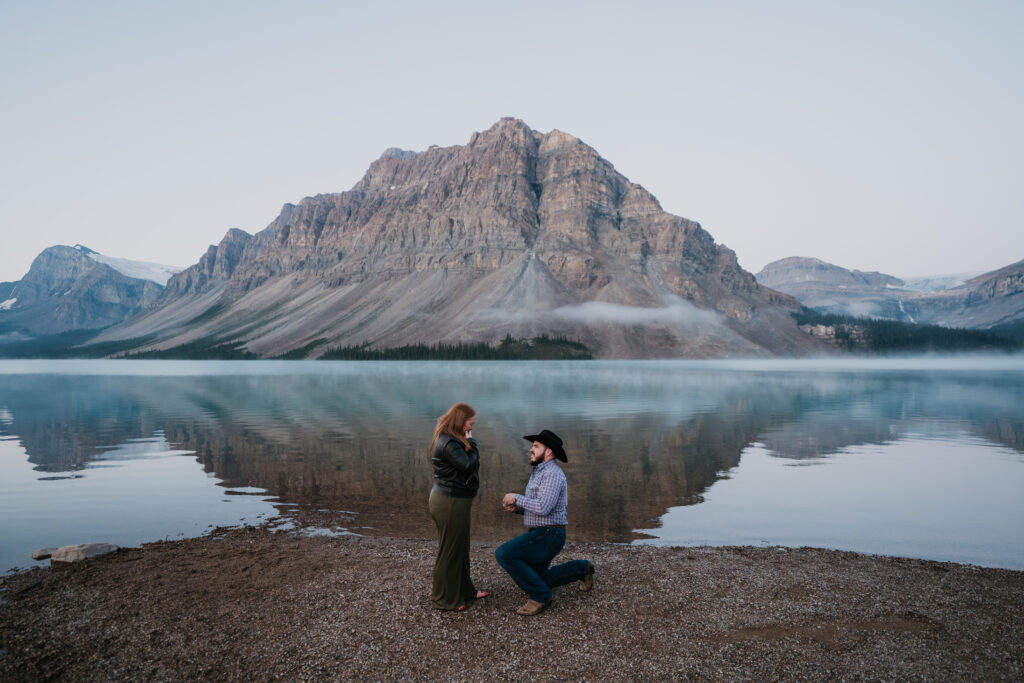 man is down on one knee in front of bow lake proposing to his girlfriend