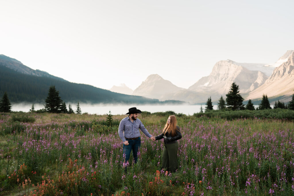 couple stands holding hands looking at each other in field of wildflowers at sunrise at bow lake