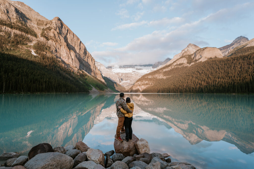couple stands on rock in front of Lake Louise as the sun comes up and reflects the mountain in the lake