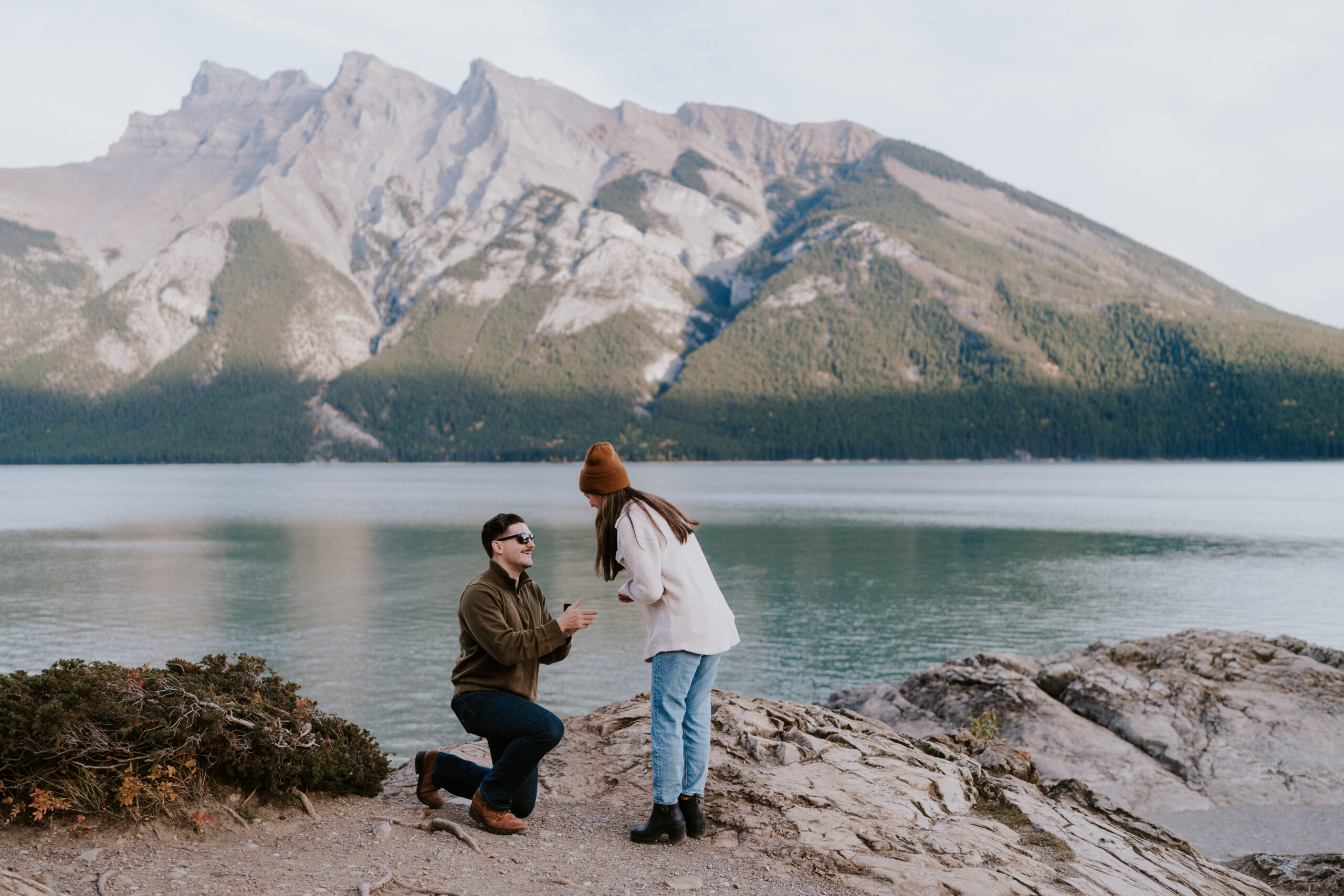 man is down on one knee proposing to his girlfriend during sunset at lake minnewanka