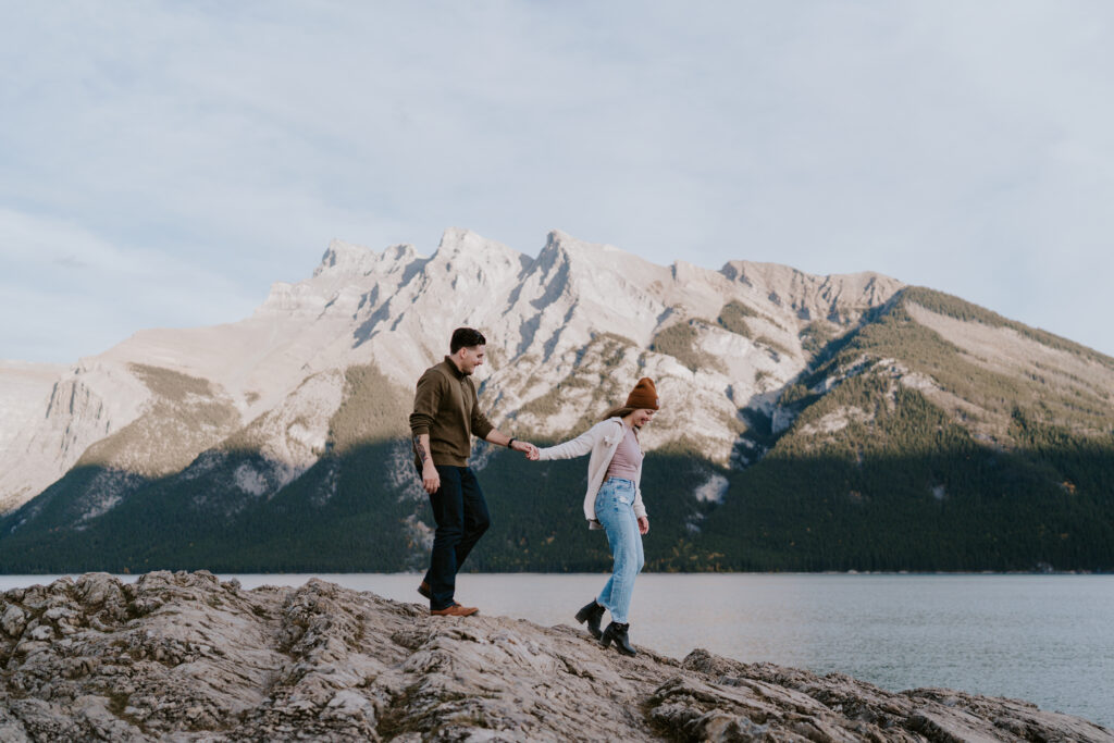 couple holds hands and walks down rocks at lake minnewanka