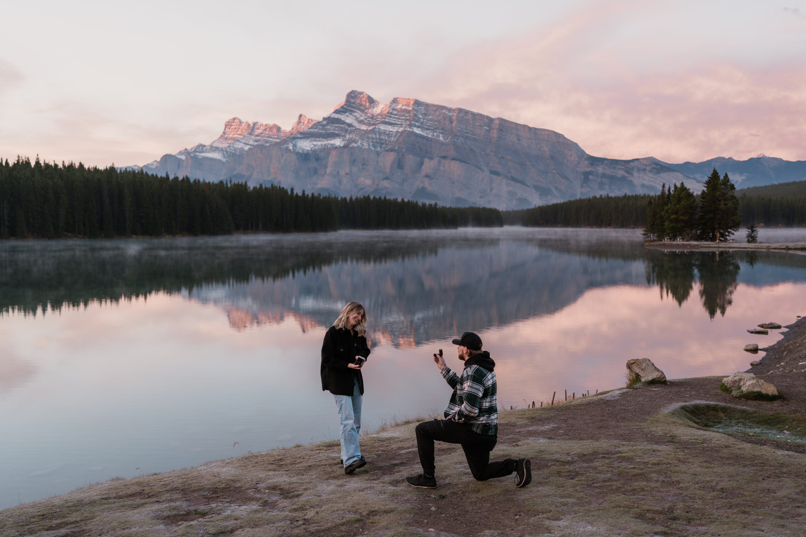 man is down on one knee proposing to his girlfriend at sunrise at two jack lake