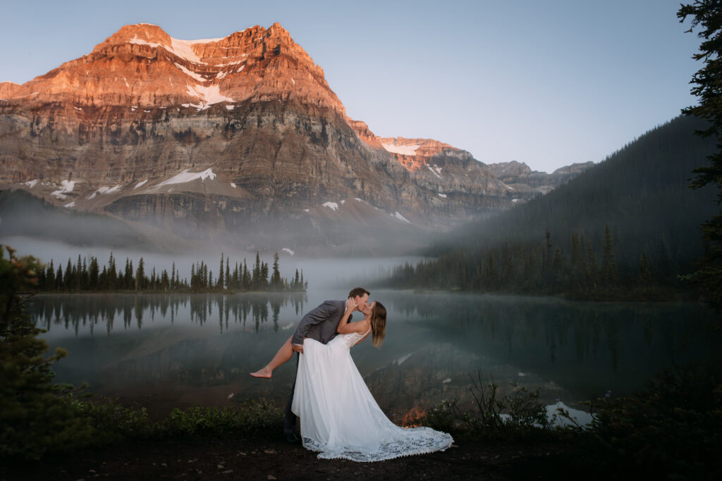 Groom dips barefoot bride at sunrise in front of Shadow Lake during elopement