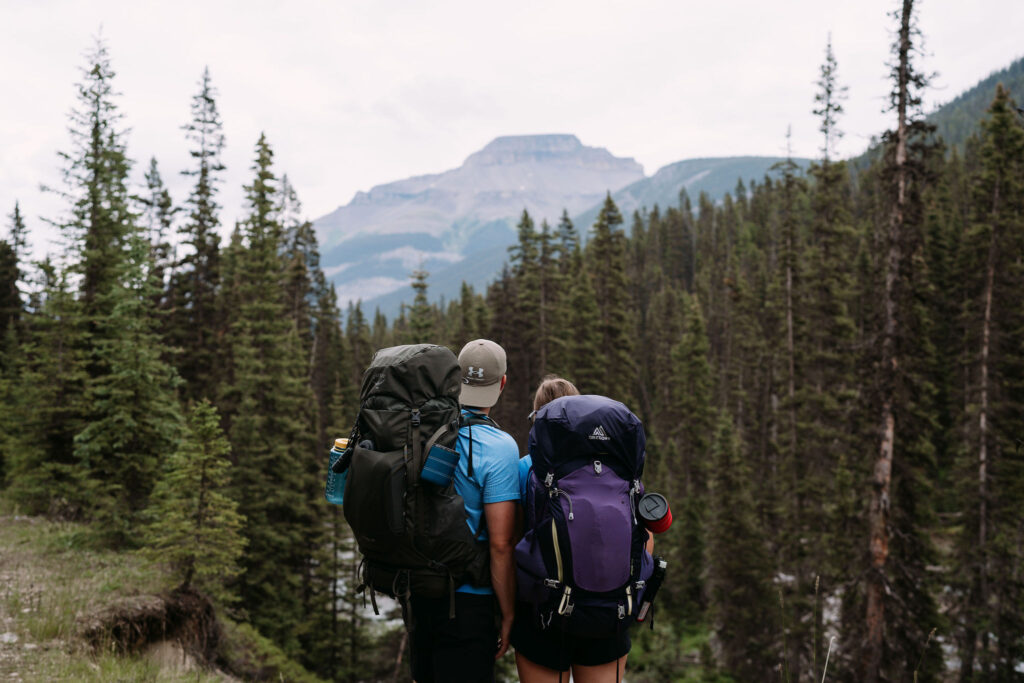 couple wearing backpack holds hands looking at mountains while hiking into Shadow Lake Lodge