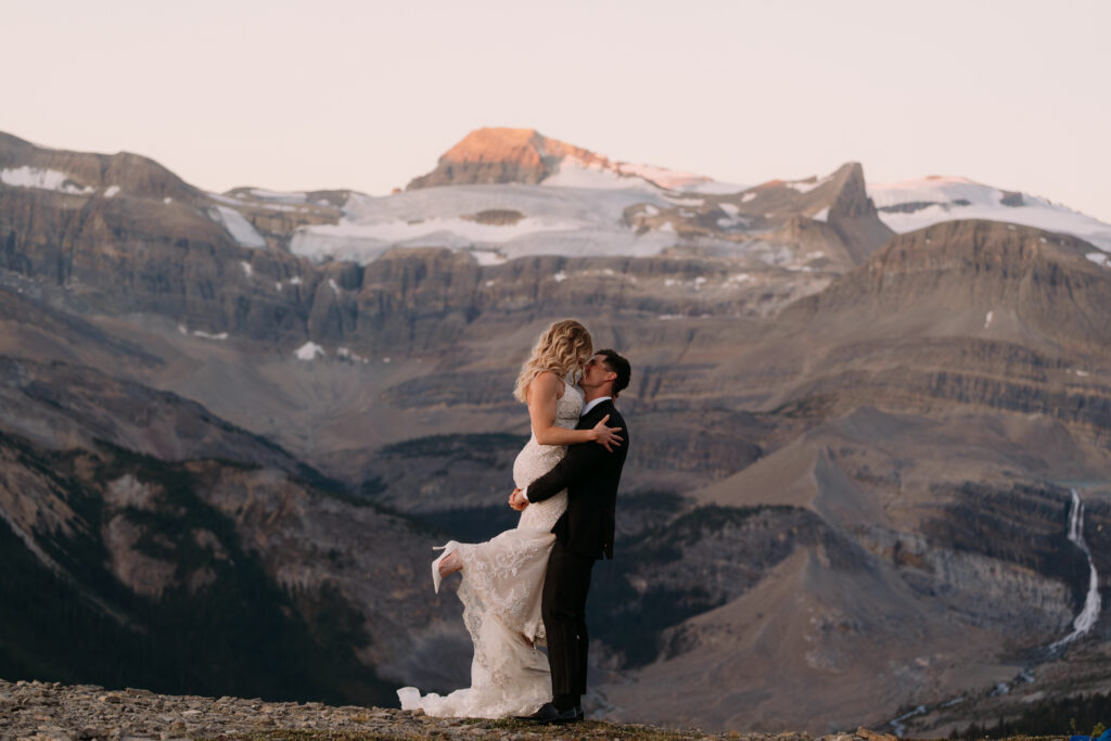groom holds bride up during sunrise mountain elopement in banff with the bride wearing white heels