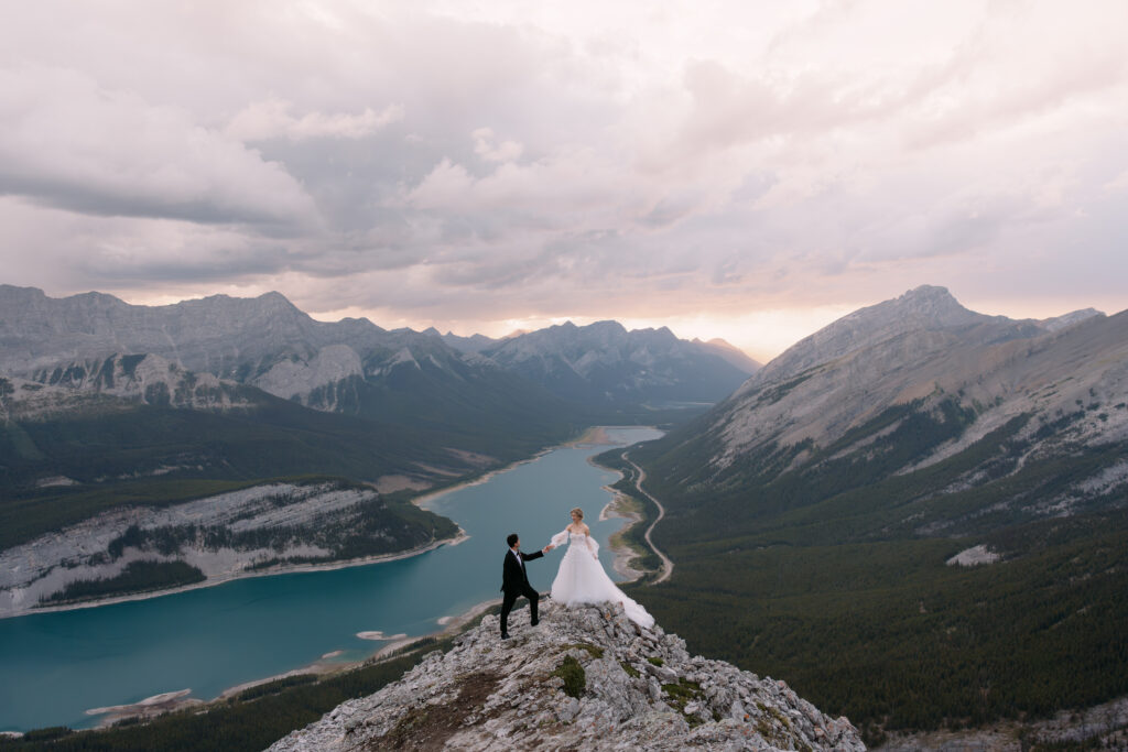 sunset hiking elopement in kananaskis bride with custom sleeves and groom stand on top of rock looking down the valley 