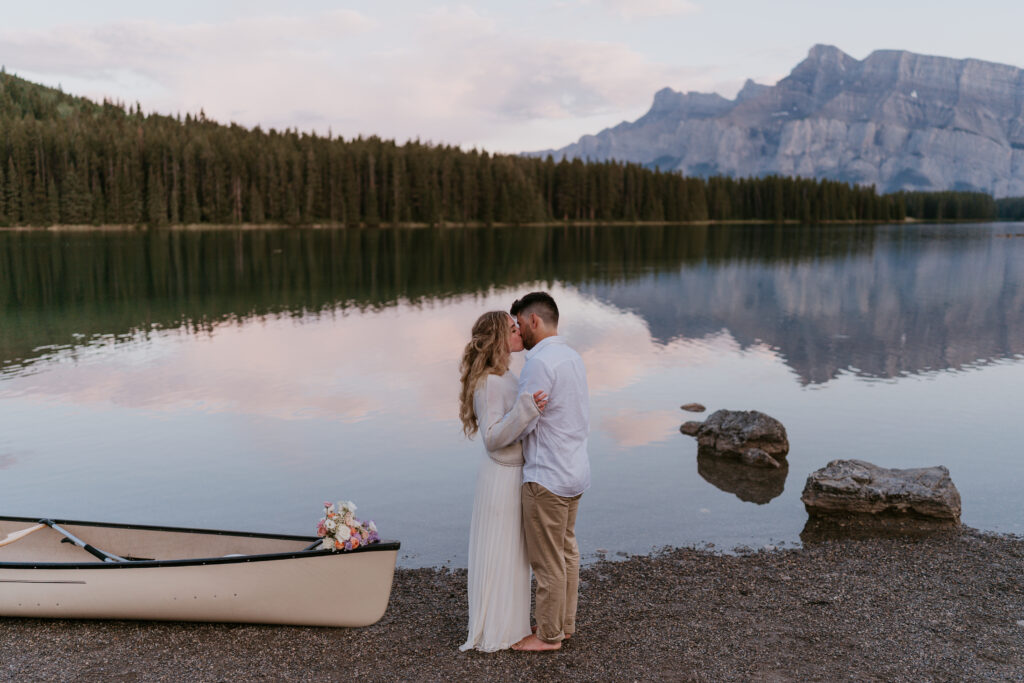 bride and groom kiss beside canoe during two jack lake elopement while bride wears a cozy sweater