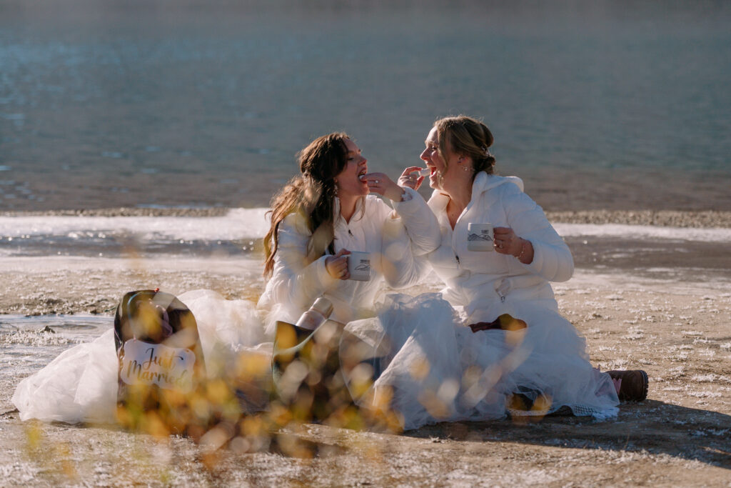 two brides with custom mugs, a just married sign, and matching white down jackets share a breakfast of powdered doughnuts