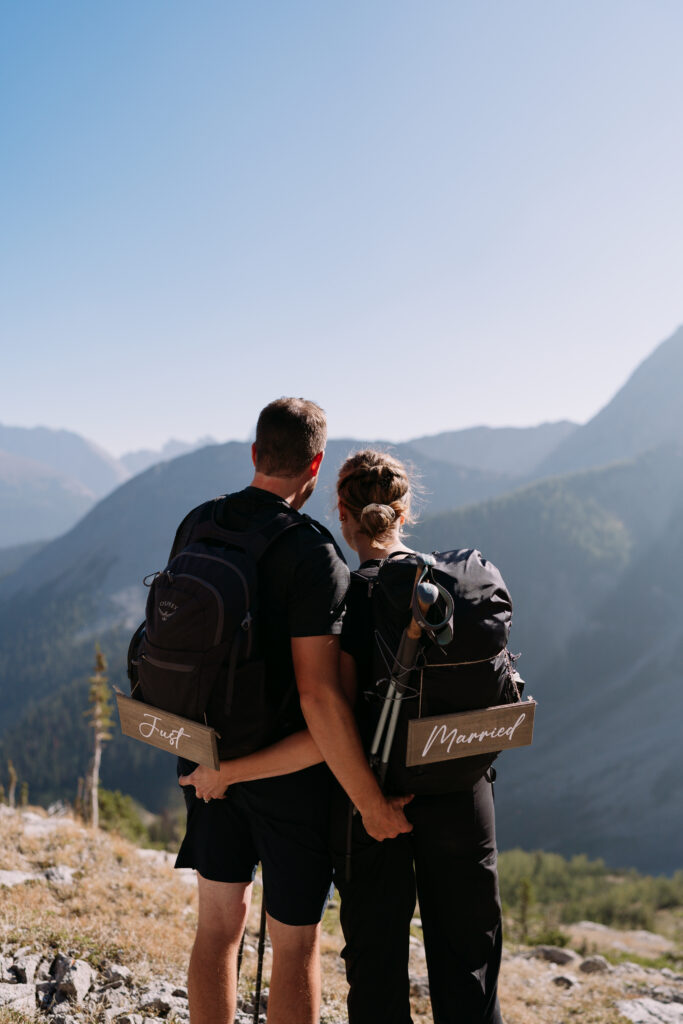 couple with hiking clothes and backpacks on look out towards the mountains with custom just married signs string onto their backpacks