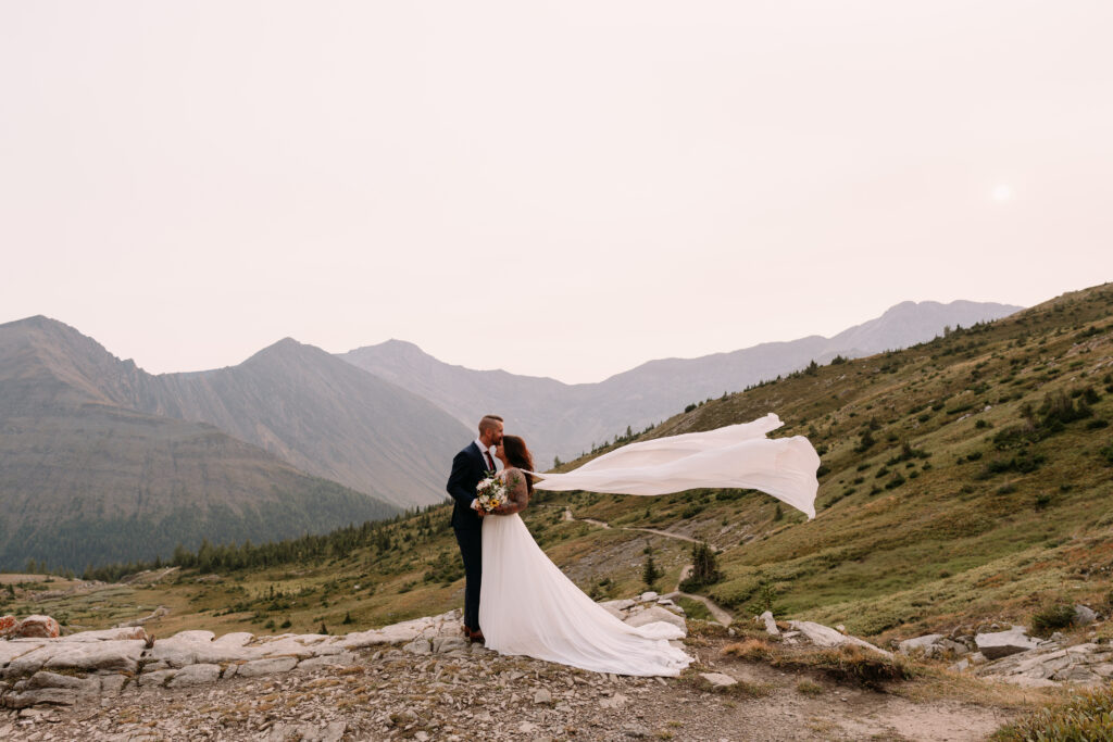 couple kisses at Ptarmigan Cirque at sunset while brides cape flows in the wind