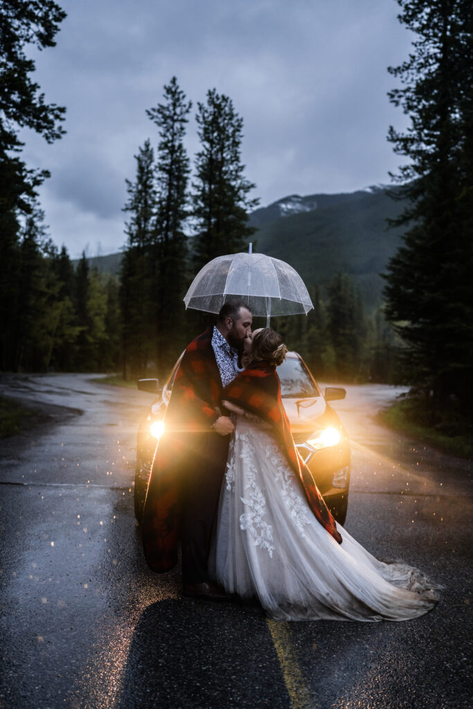 couple kisses wrapped in a red blanket under an umbrella with the glow of headlights from their car behind them 