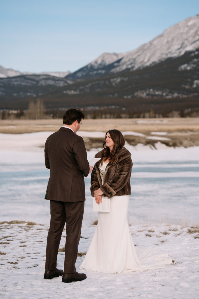 bride and groom share vows on the snow during nordegg elopement while bride wears a short brown mink coat overtop dress 
