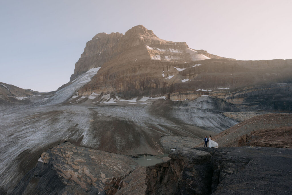 couple on cliff holds hands during sunset helicopter elopement in front of glacier
