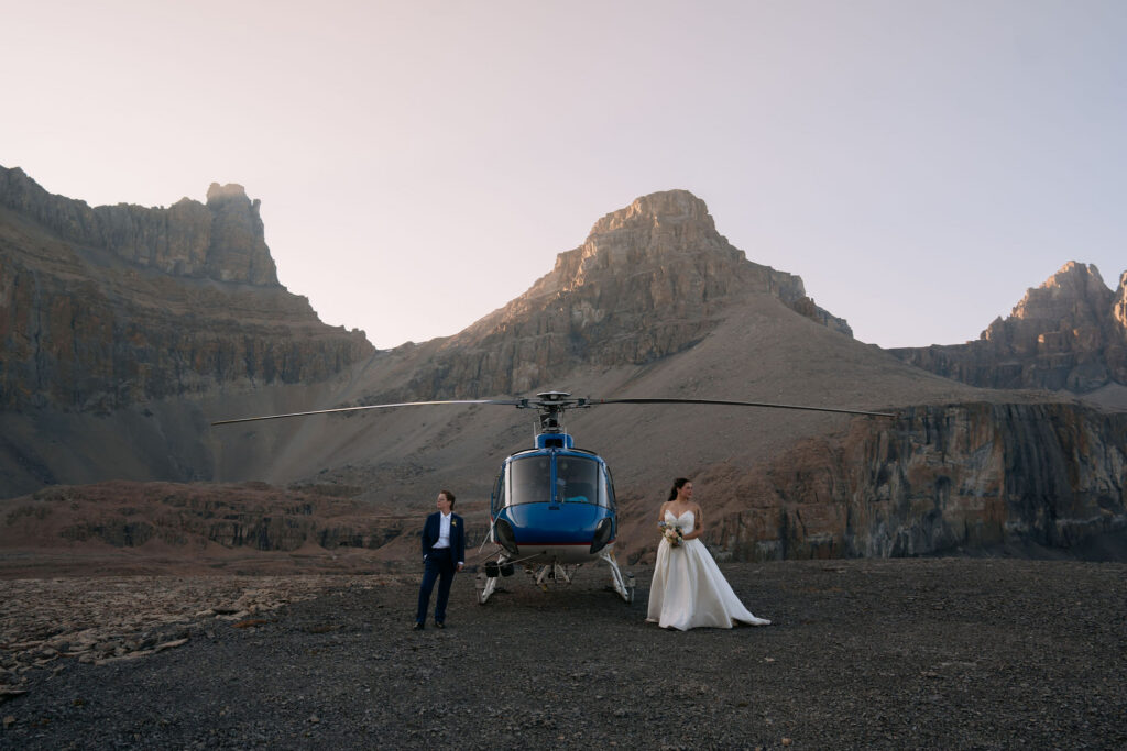 brides stand on either side of a blue helicopter during elopement