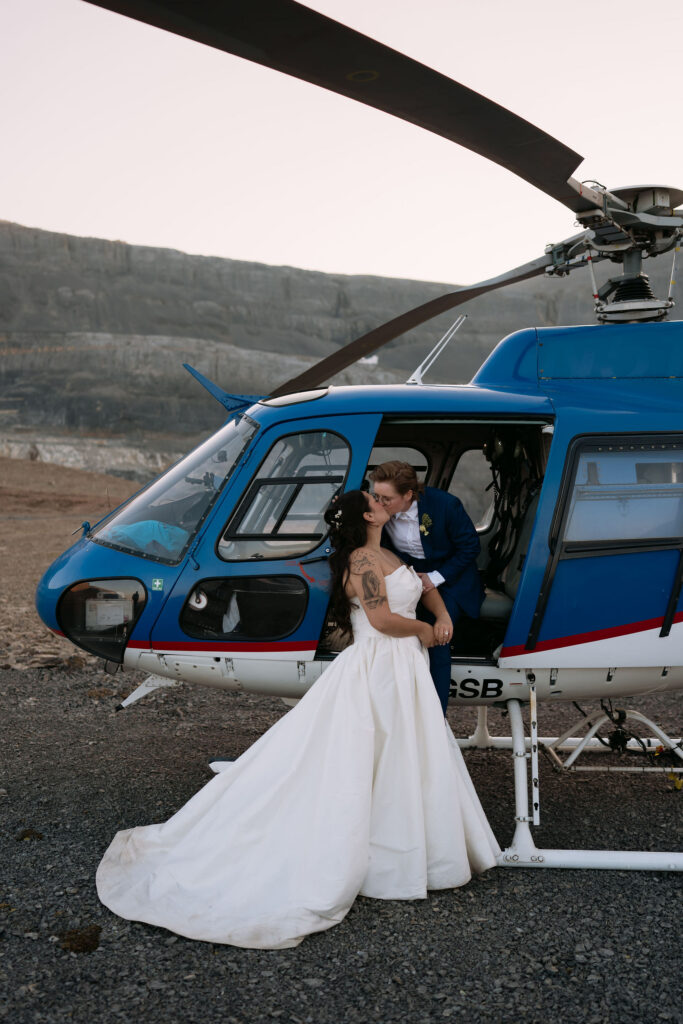 bride in blue suit sits in blue helicopter door kissing bride in white gown standing in front of her