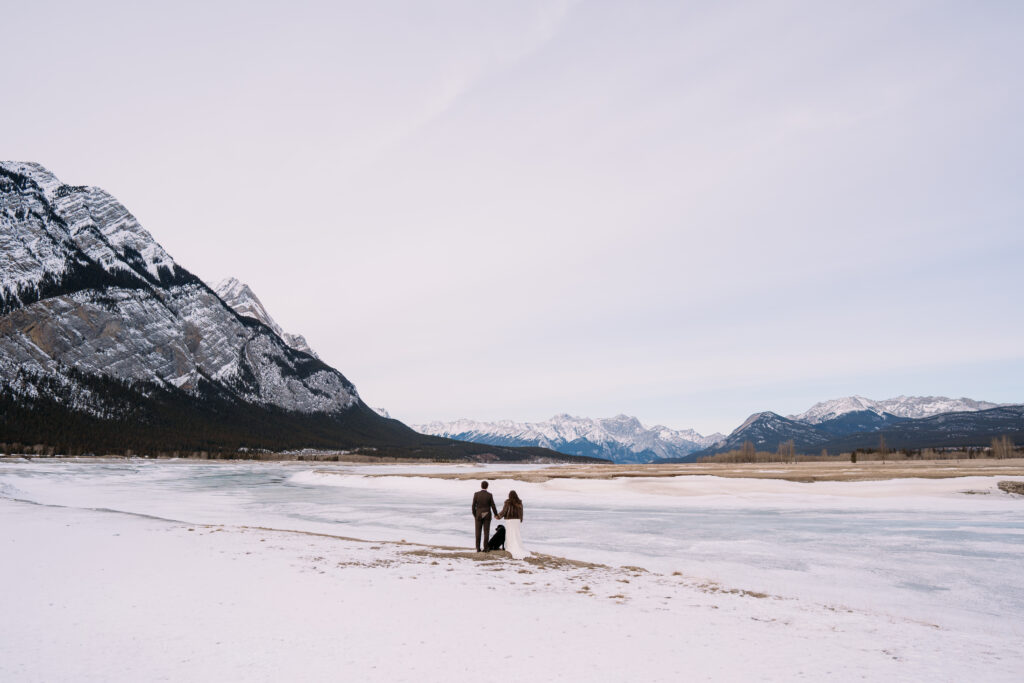 Bride and groom hold hands with black dog in between them on the banks of the river during winter Nordegg elopement