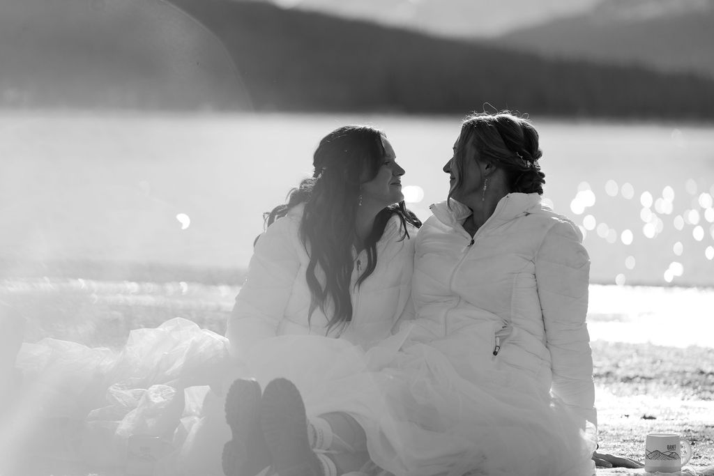 brides in white down jackets look at each other during bow lake elopement