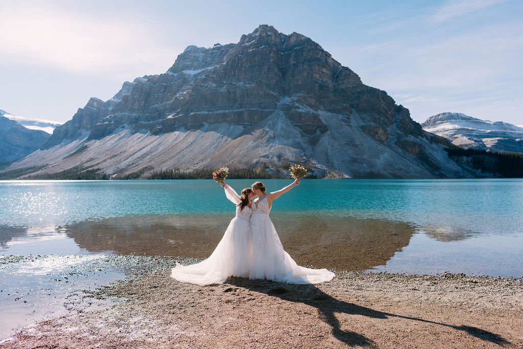 Brides hold up bouquets and kiss at Bow Lake
