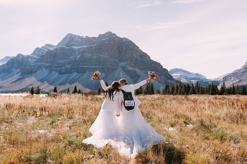 brides hold up bouquets kissing while wearing a backpack with just married sign at Bow Lake