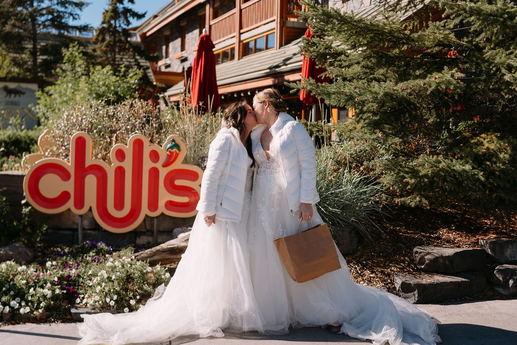 Brides kiss outside Chili's Banff in wedding dresses and white down jackets.