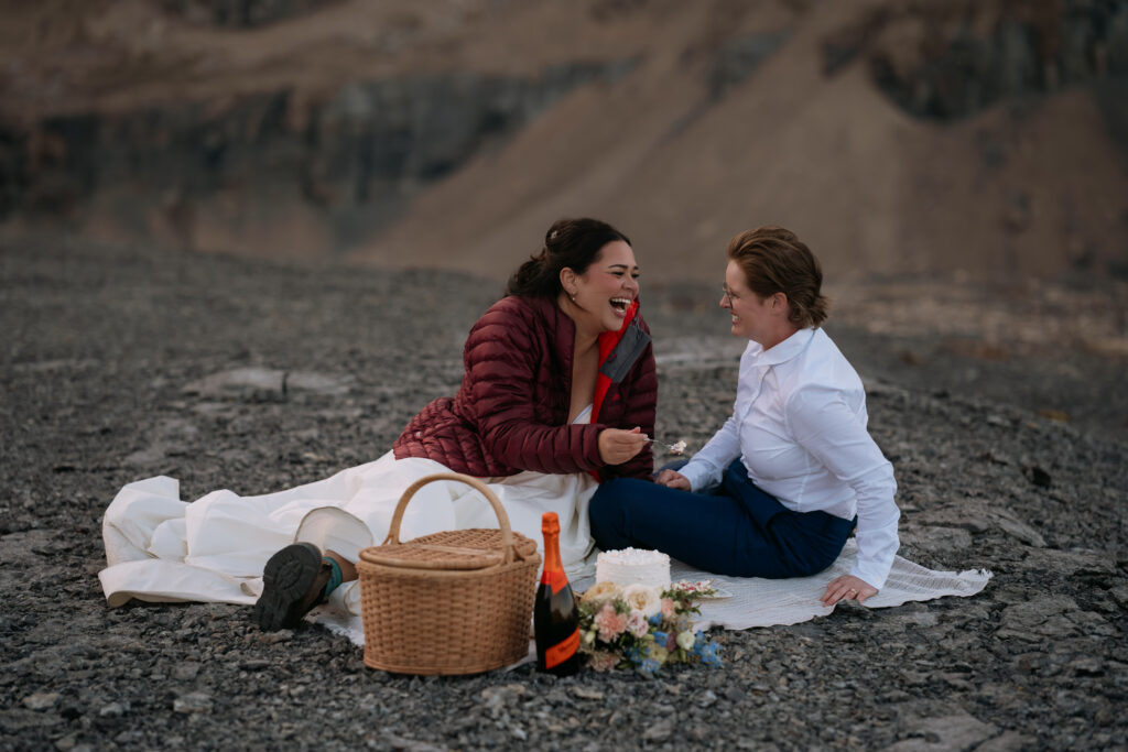 couple shares picnic on rocks with picnic basket, cake, wine, during helicopter elopement